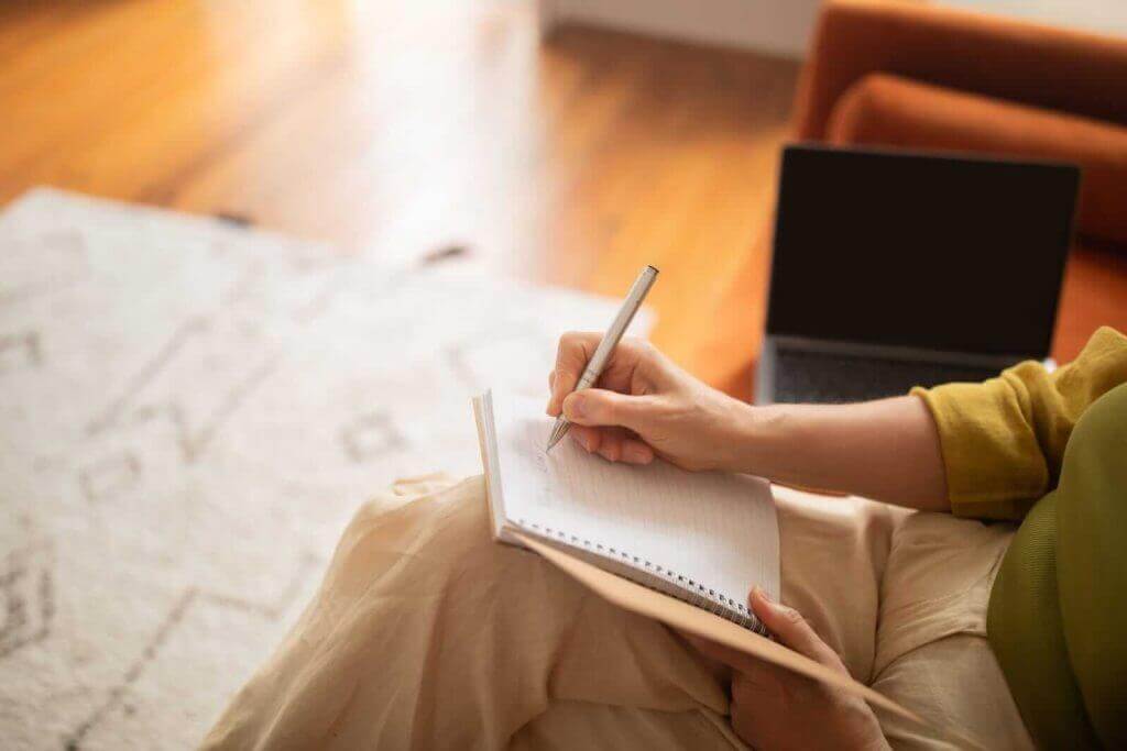 A person sitting on the floor writing in a notebook with a laptop nearby