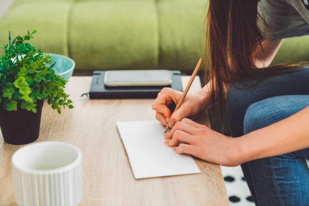 A person seated at a table writing on a notepad with a pencil with a potted plant cup and smartphone nearby