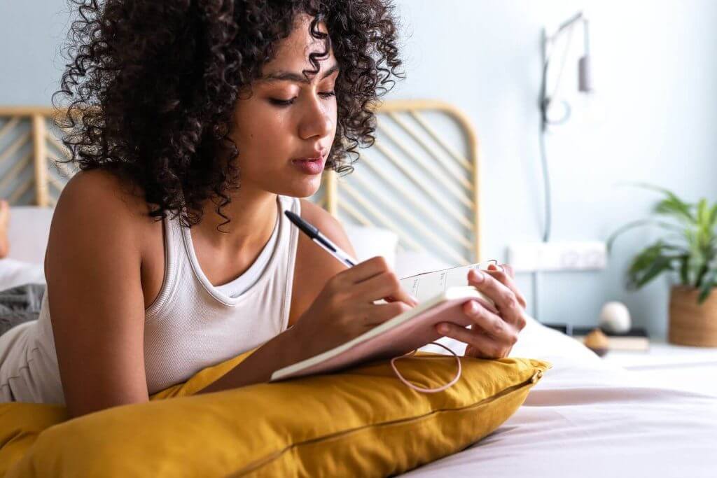 A woman writing in a notebook while reclining on a bed with a yellow pillow