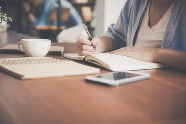 Person writing in notebook with coffee cup and smartphone on table