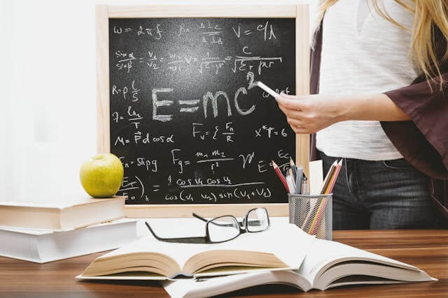 A person standing beside a chalkboard filled with mathematical equations pointing to the equation e=mc² with an open book eyeglasses and an apple on the desk