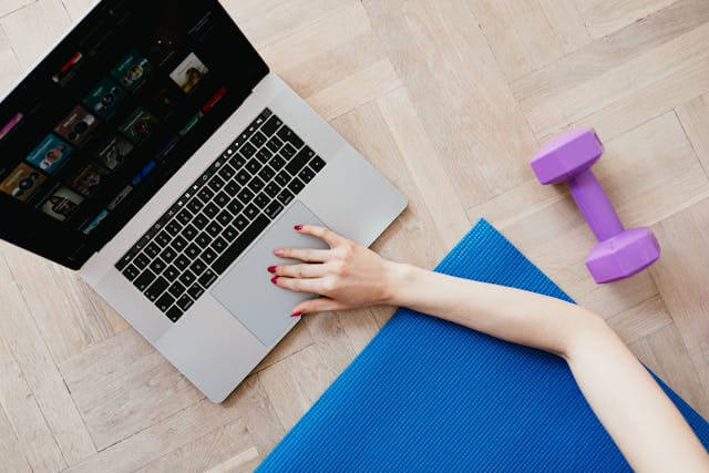 Person lying on a yoga mat beside a laptop and dumbbells possibly following an online workout