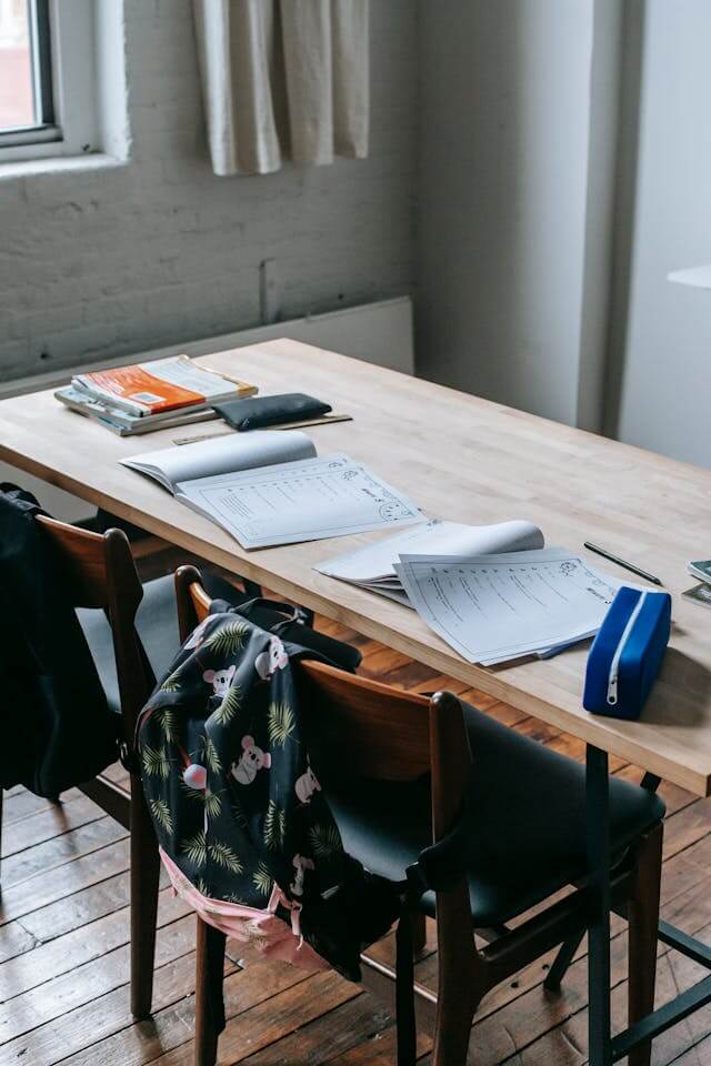 A wooden table with books notebooks and a backpack in a room with natural light