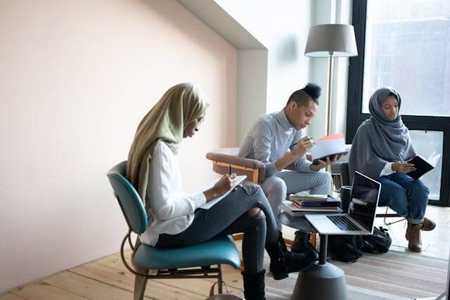 Three people sit in a well lit room engaged in individual tasks with notepads and laptops