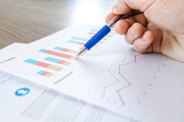 Hand holding a pen pointing at bar and line graphs on printed charts analyzing data on paper