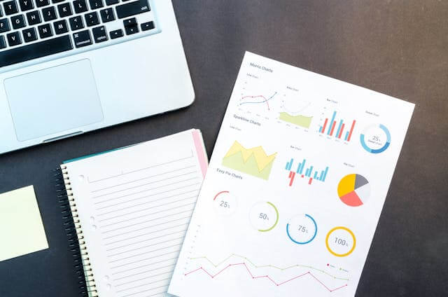 A laptop a notepad a sheet with various colorful charts and graphs and a sticky note are displayed on a desk