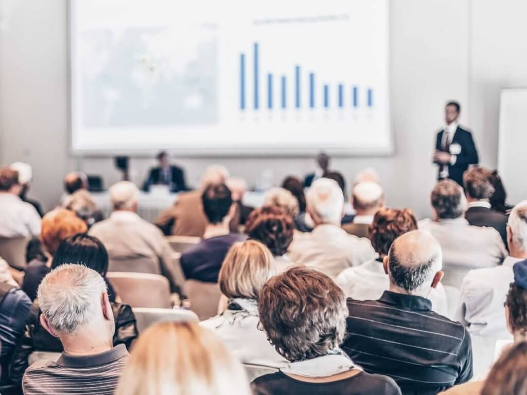 A speaker presents a graph during a business conference to an audience seated in a dimly lit conference room