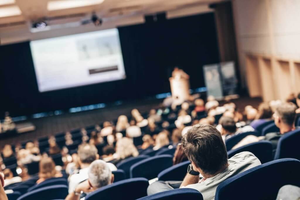 A conference room filled with people seated in rows of blue chairs focusing on a presenter at a podium near a screen displaying a presentation