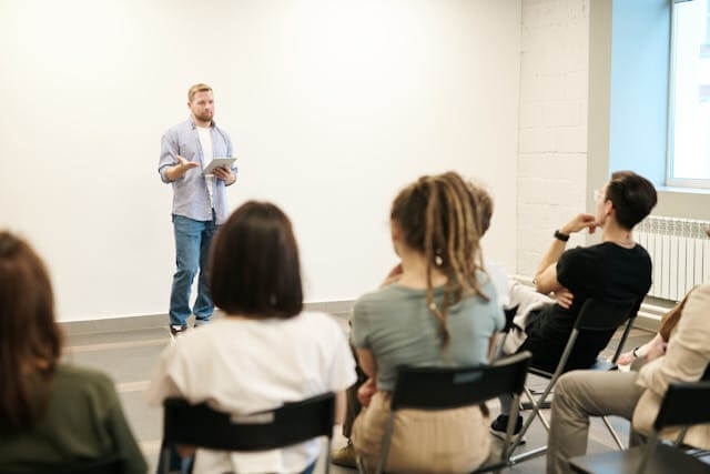 Person gives a standing presentation holding a tablet in front of a seated audience in a brightly lit room