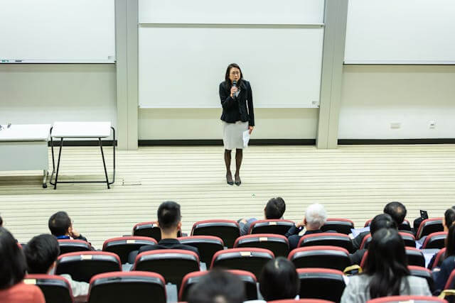 A woman stands at the front of a lecture hall speaking into a microphone People are seated in rows facing her listening attentively A whiteboard is visible in the background