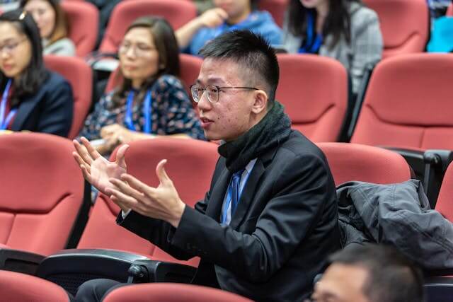 A man in a suit and scarf is seated in a red auditorium gesturing with both hands while speaking Other people are seated around him appearing attentive