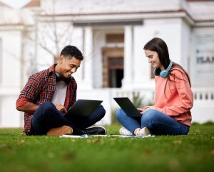 Two people sitting on grass, using laptops and wearing headphones, with a building in the background.