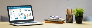 A laptop displaying website traffic analytics sits on a desk next to sunglasses, notebooks, a potted plant, and a cup holding pens and pencils.