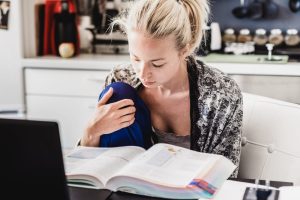 A woman with a ponytail studies by reading a book at a desk, with a laptop open nearby. She is sitting in a kitchen.