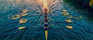 aerial view of a rowing team in a long boat, paddling in sync on a calm, golden-lit water body.