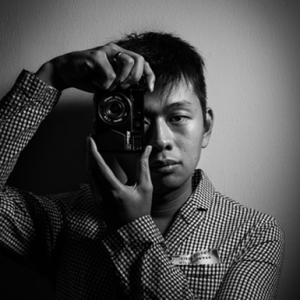 black and white photo of a person holding a camera up to their face, partially obscuring one eye. the person is wearing a checkered shirt and looking directly at the camera.