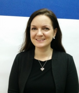 A woman in a black jacket standing in front of a blue wall.